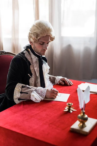 Selective focus of handsome victorian man in wig holding feathers and writing letter — Stock Photo