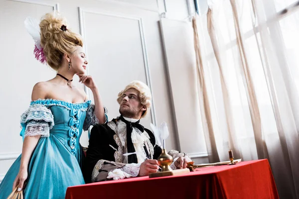 Low angle view of victorian girl in wig standing near handsome pompous man holding feather near paper — Stock Photo