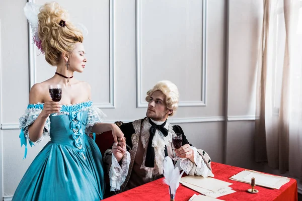 Victoriana chica en peluca cogido de la mano con guapo pomposo hombre con vidrio vino sentado cerca de papeles - foto de stock