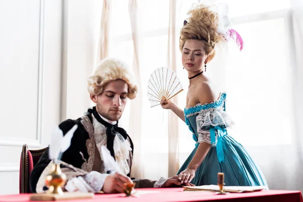 Selective focus of victorian woman in wig holding fan near handsome man writing letter — Stock Photo