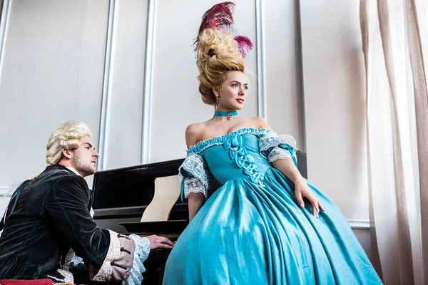 Attraktive viktorianische Frau im blauen Kleid steht neben einem Mann mit Perücke und spielt Klavier — Stockfoto