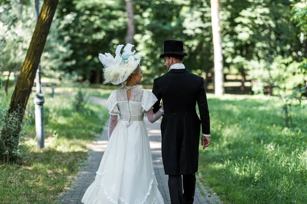 Victorian man and woman in hats walking outside near green trees — Stock Photo