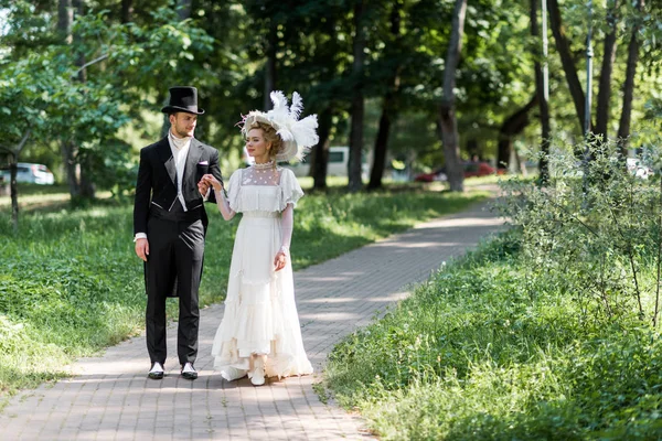 Schöner viktorianischer Mann und attraktive Frau mit Hüten halten Händchen, während sie draußen spazieren gehen — Stockfoto