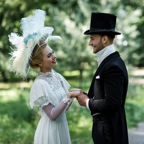 Cheerful victorian man and woman in hats holding hands while standing outside — Stock Photo