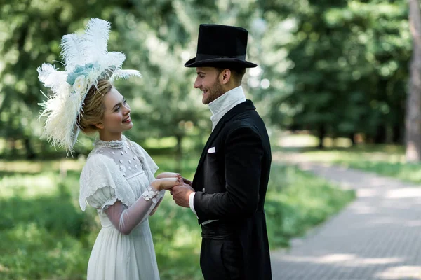 Glückliche junge viktorianische Frau und gutaussehender Mann mit Hut, Händchen haltend draußen — Stockfoto