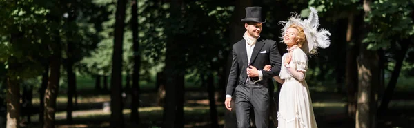 Panoramic shot of happy young victorian woman and handsome man in hat walking outside — Stock Photo