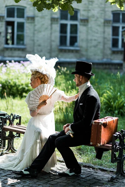 Atractiva mujer victoriana sosteniendo ventilador mientras está sentado con un hombre guapo en el banco - foto de stock