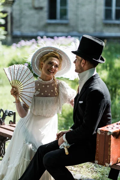 Happy victorian woman holding fan while sitting with handsome man on bench — Stock Photo