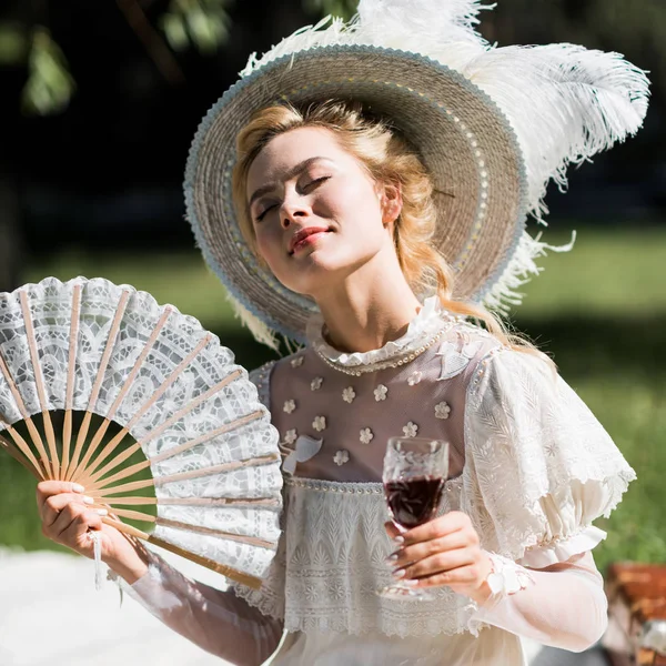 Alegre joven victoriana sosteniendo ventilador y copa de vino - foto de stock