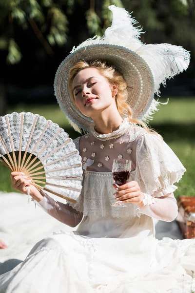 Cheerful young victorian woman with closed eyes holding fan and wine glass — Stock Photo