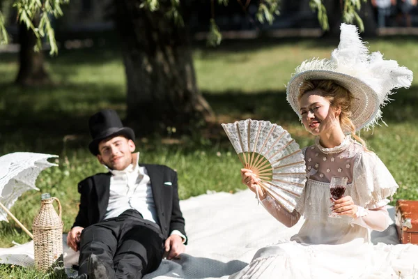 Enfoque selectivo de la mujer victoriana feliz sentado en manta y sosteniendo copa de vino cerca del hombre - foto de stock