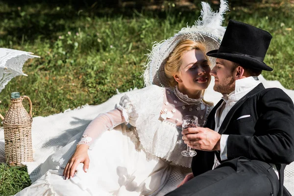 Mujer victoriana feliz sentado en la manta y mirando al hombre en sombrero sosteniendo copa de vino - foto de stock