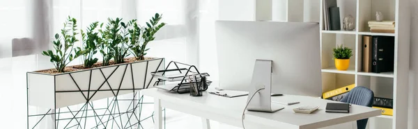 Panoramic shot of table with computer and document tray near bookcase and flowerpot with plants — Stock Photo