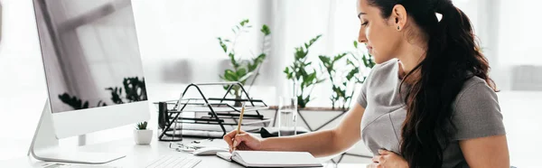 Panoramic shot of pregnant woman sitting behind table with computer, keyboard and document tray, writing notes and holding belly — Stock Photo