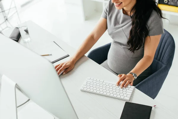 Abgeschnittene Ansicht einer schwangeren Frau, die hinter einem Tisch sitzt und am Computer arbeitet — Stockfoto