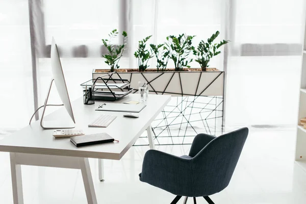 Table blanche avec ordinateur, plateau de documents, verre et ordinateur portable près de la chaise de bureau et pot de fleurs avec plante — Photo de stock