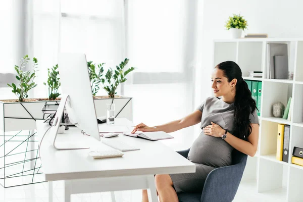 Pregnant woman sitting in room with bookcase and flowerpot with plants behind table and working on computer — Stock Photo