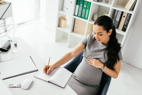 Vista ad alto angolo di donna incinta prendere appunti nel notebook mentre seduto dietro il tavolo vicino alla libreria — Foto stock