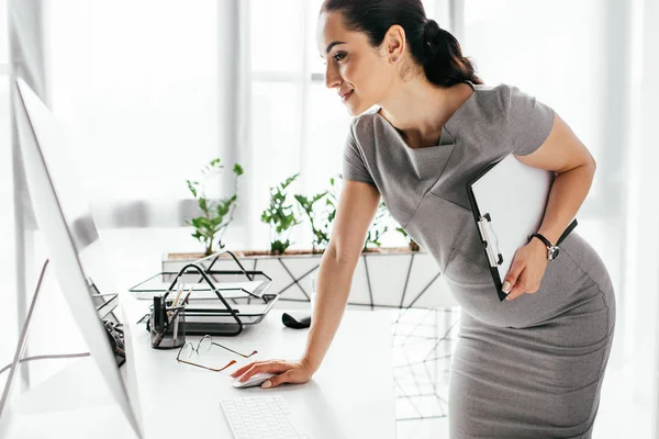 Pregnant woman standing near table, holding clipboard and working on computer — Stock Photo