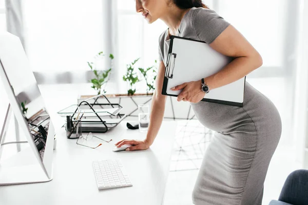 Vista cortada da mulher grávida em pé perto da mesa, segurando pasta e trabalhando no computador — Fotografia de Stock