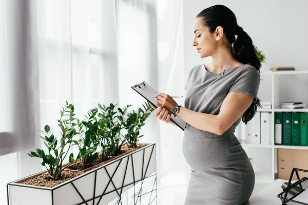 Seitenansicht einer schwangeren Frau, die im Büro Notizen in Notizbuch macht — Stockfoto