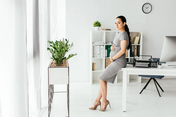 Vue pleine longueur de la femme enceinte debout au bureau avec pot de fleurs avec plante, table et bibliothèque — Photo de stock