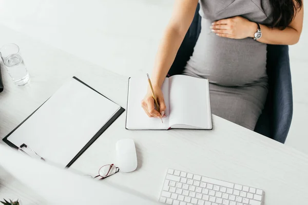 Cropped view of pregnant woman taking notes in notebook while sitting behind table with keyboard, folder and glass with water in office — Stock Photo