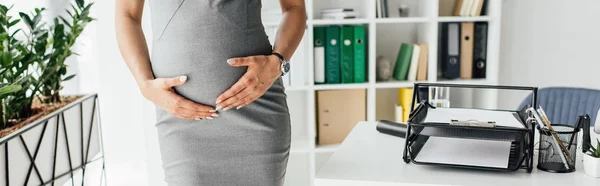 Vue recadrée de la femme enceinte tenant ventre tout en restant au bureau avec pot de fleurs et bibliothèque près de la table avec plateau de documents — Photo de stock