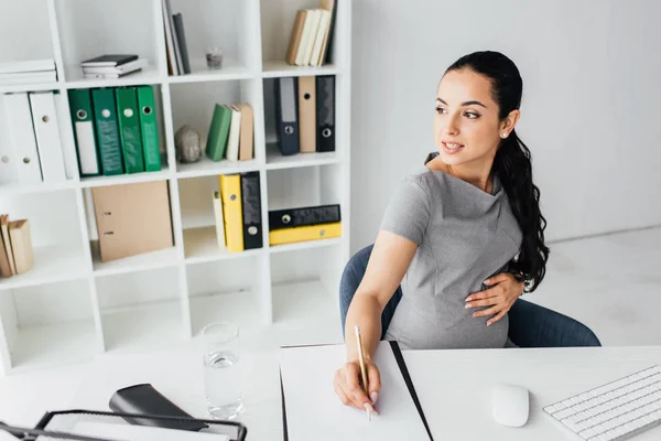 Pregnant woman taking notes while sitting behind table in office and looking away — Stock Photo
