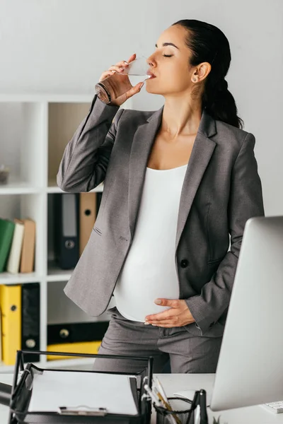 Femme enceinte en costume debout dans le bureau près de la table avec ordinateur et plateau de documents et d'eau potable — Photo de stock