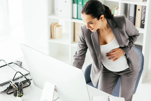 Schwangere im Anzug hält Bauch, während sie am Tisch steht und am Computer arbeitet — Stockfoto