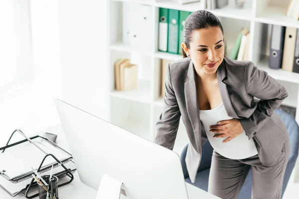 Femme enceinte en costume tenant ventre tout en se tenant près de la table, travaillant sur l'ordinateur et regardant loin — Photo de stock