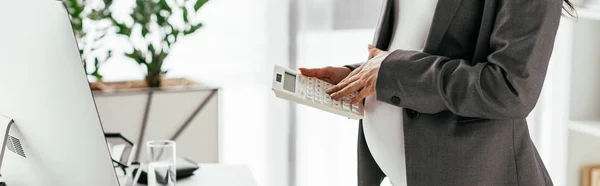 Panoramic shot of pregnant woman calculating while standing near table with computer and notebook in office — Stock Photo