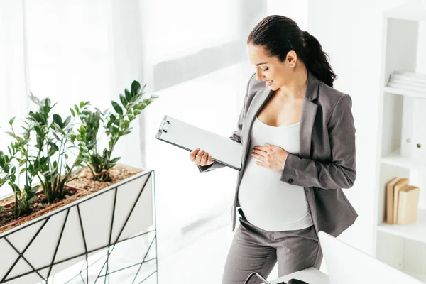 Vista de ángulo alto de la mujer embarazada mirando a la carpeta mientras está de pie en la oficina cerca de maceta con planta - foto de stock