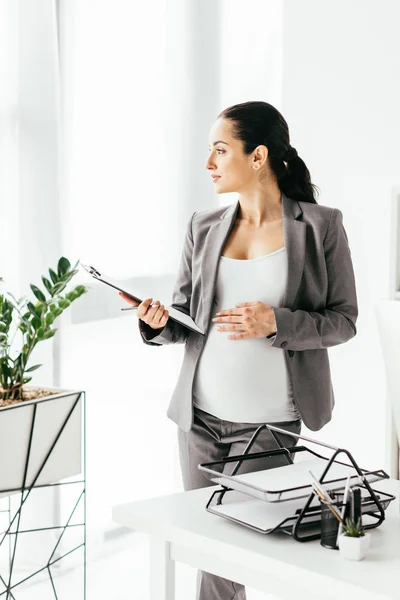 Pregnant woman holding belly and folder while standing in office near flowerpot and table with document tray and looking away — Stock Photo