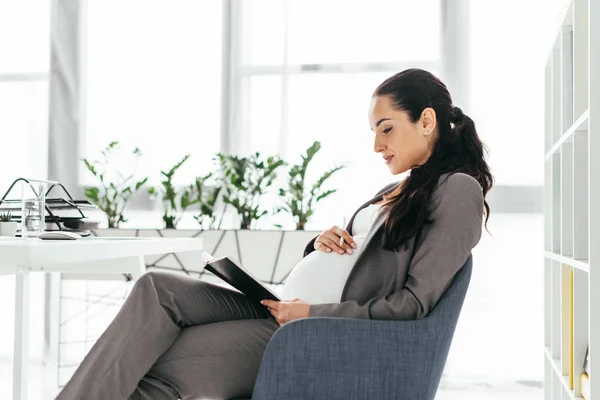 Pregnant woman sitting in office in office chair and reading — Stock Photo