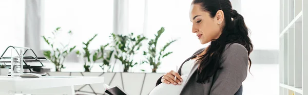 Tiro panorâmico de mulher grávida sentada no escritório em cadeira de escritório e leitura — Fotografia de Stock