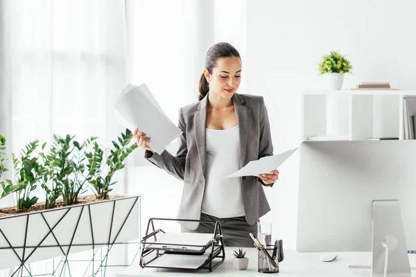 Mujer embarazada leyendo papeles mientras está de pie en la oficina cerca de maceta y mesa con computadora, bandeja de documentos y caja de lápiz - foto de stock