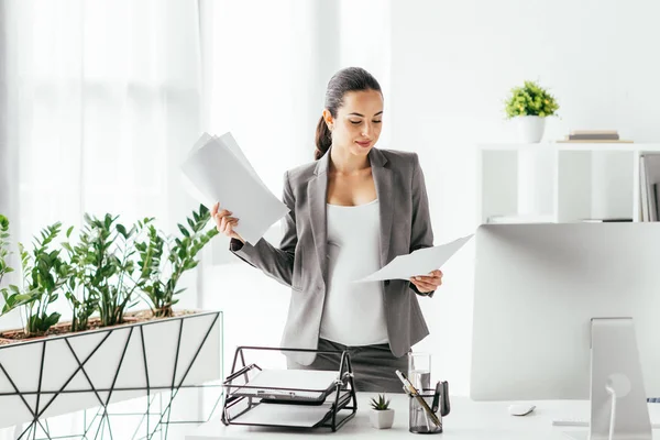 Femme enceinte lisant des papiers tout en restant au bureau près du pot de fleurs et de la table avec ordinateur, plateau de documents et boîte à crayons — Photo de stock