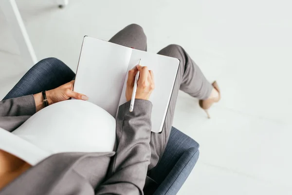 Vista ritagliata donna incinta prendere appunti nel notebook mentre seduto in ufficio in sedia ufficio — Foto stock