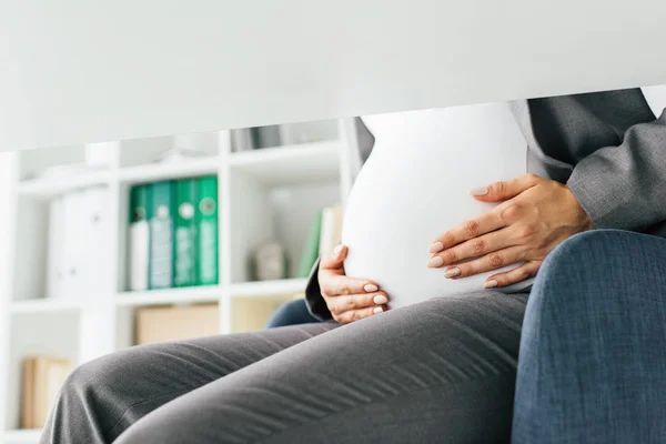 Vista cortada da mulher grávida segurando barriga enquanto sentado na cadeira do escritório — Fotografia de Stock