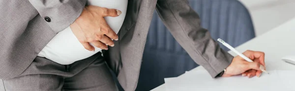 Panoramic shot of pregnant woman in suit holding belly and making notes in notebook — Stock Photo