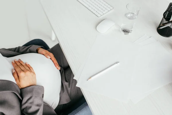 Cropped view of pregnant woman sitting behind table with glass, keyboard, papers and pencil — Stock Photo