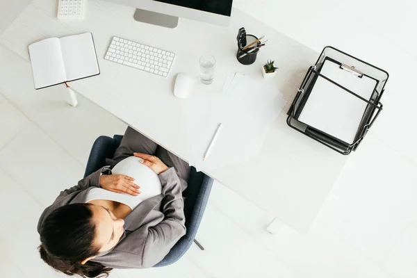 Top view of pregnant woman sitting behind table with computer, notebook, document tray, calculator, glass, keyboard, papers and pencil — Stock Photo