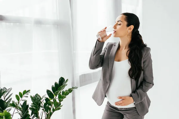 Pregnant woman drinking water whie standing in office near plant — Stock Photo