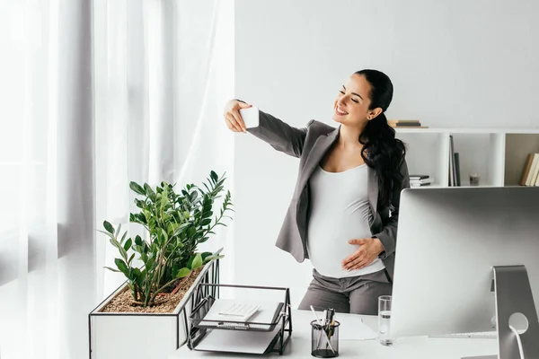 Femme enceinte debout près de la table et pot de fleurs, et de prendre selfie — Photo de stock