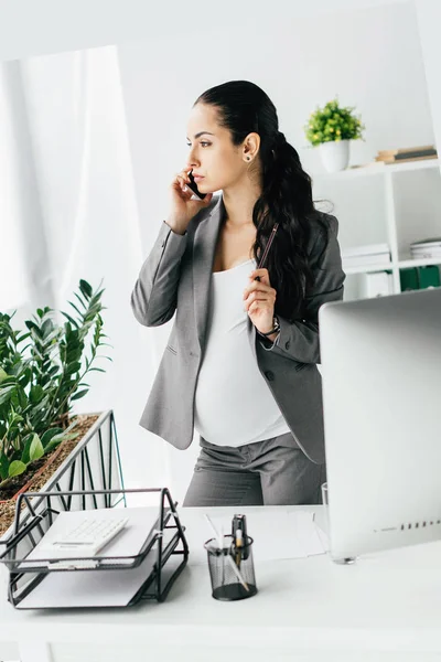 Pregnant woman standing near table in office, talking on smartphone and looking away — Stock Photo