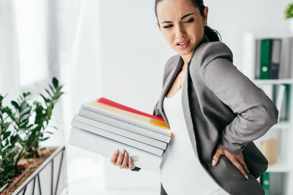 Pregnant woman enduring pain and holding folders while standing in office — Stock Photo