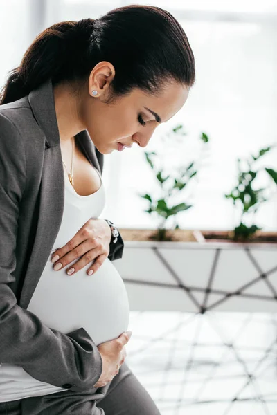 Tense pregnant woman holding and looking at belly while standing in office near flowerpot — Stock Photo