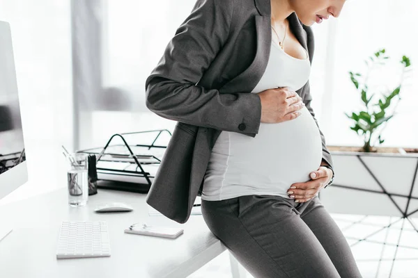 Cropped view of tense pregnant woman holding and looking at belly while standing in office near flowerpot — Stock Photo